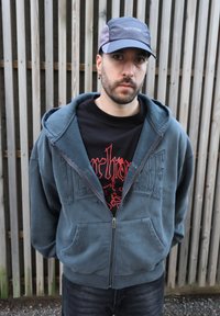 Sweat à capuche zippé doublé en polaire bleu foncé avec poches avant et détails brodés, porté sur un t-shirt noir avec un design graphique rouge.