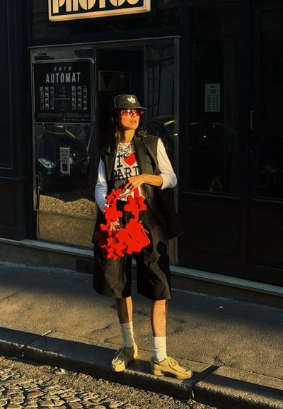 Chaleco negro sin mangas sobre camisa blanca de manga larga y camiseta gráfica, pantalones cortos negros, zapatos amarillos, bolso rojo con forma de flor y gafas de sol.