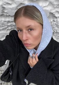 Donna con capelli biondi che indossa un felpa grigia sotto un cappotto nero, tiene il felpa vicino al mento, in piedi contro un muro bianco testurizzato.