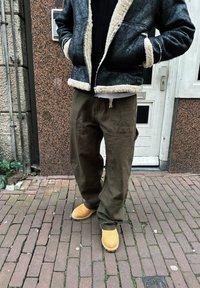 Veste en peau de mouton noire doublée de fourrure, pantalons vert olive et bottes en suede beige. Matériaux texturés avec une coupe décontractée. Détail de fermeture éclair visible sur la veste.