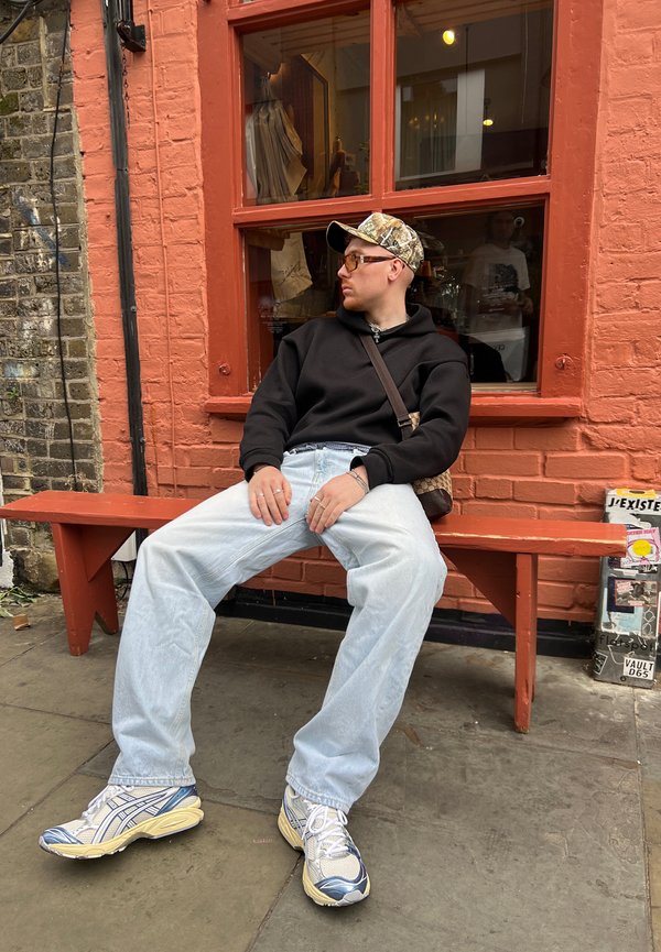 Persona sentada en un banco rojo, vistiendo una sudadera negra, jeans de mezclilla azul claros y una gorra con estampado. Lleva gafas de sol metálicas y zapatillas gruesas.