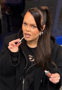 Brun læbestift med sølvdetaljer holdt af en person, der påfører læbestift. Sort hættetrøje med udskæringsdetaljer, langt, mørkt hår.