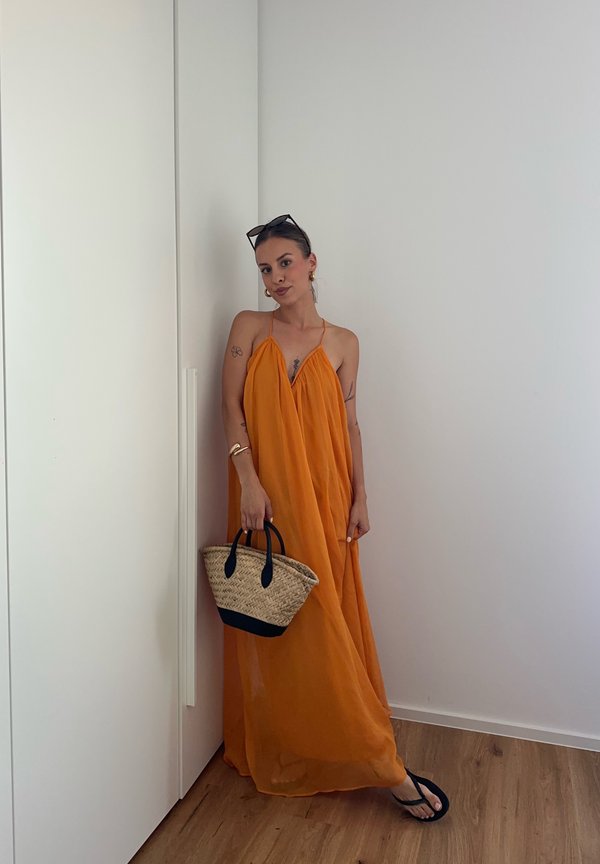 Mujer con vestido naranja sin mangas sosteniendo un bolso tejido, de pie en interior contra una pared blanca y suelo de madera, con gafas de sol y sandalias.
