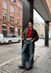 Femme en polo rouge et veste verte, appuyée contre un pilier noir sur un trottoir de la ville, avec des bâtiments en brique et colorés en arrière-plan.