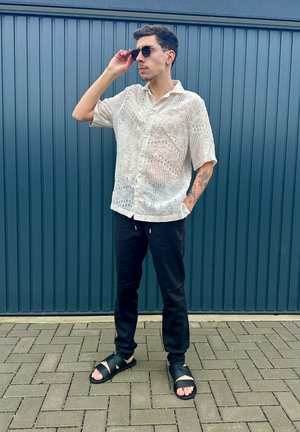 Camisa blanca de manga corta con estampado, pantalones negros y sandalias negras. La camisa presenta un diseño texturizado con botones y mangas cortas.