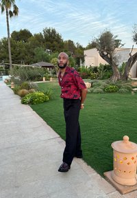 Kortärmad röd skjorta med svart blommönster, svarta byxor och mörka skor; står på en stenslagd väg bredvid grön gräs och växter.