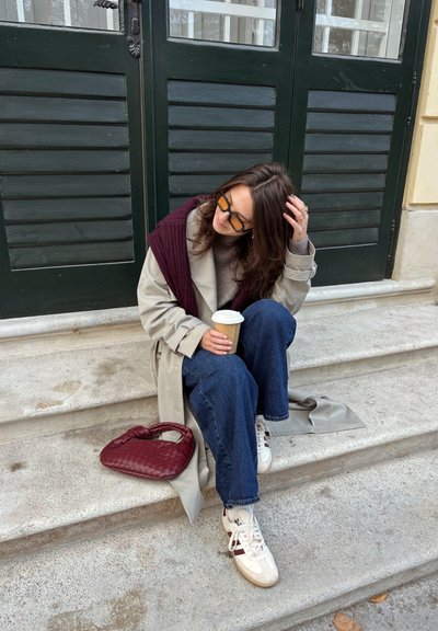 Abrigo trench beige sobre un suéter gris, combinado con vaqueros de pierna ancha y zapatillas blancas. Sosteniendo un vaso de café de papel, al lado un bolso burdeos.