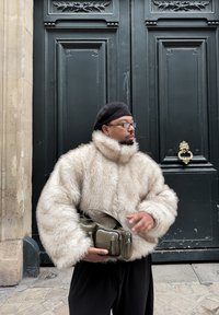 Manteau en fausse fourrure beige clair avec un col montant et des manches surdimensionnées, accompagné d'un petit sac vert olive avec plusieurs poches à fermeture éclair.