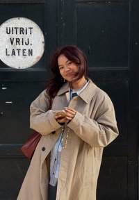 Beige rutete overdimensjonert jakke, stripete skjorte under, holder en telefon, med en rød lærveske. Mørkegrønn vegg med et rundt hvitt skilt.