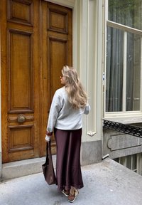 Szary sweter, bordowa spódnica, brązowa torba; stoi przy dużych drewnianych drzwiach. Gładkie tekstury, miękkie tkaniny, casualowy design i stonowane kolory.