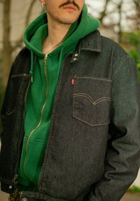 Veste en denim foncé avec surpiqûres orange, dotée de deux poches poitrine. Portée par-dessus un sweat à capuche vert zippé avec de grandes poches kangourou.