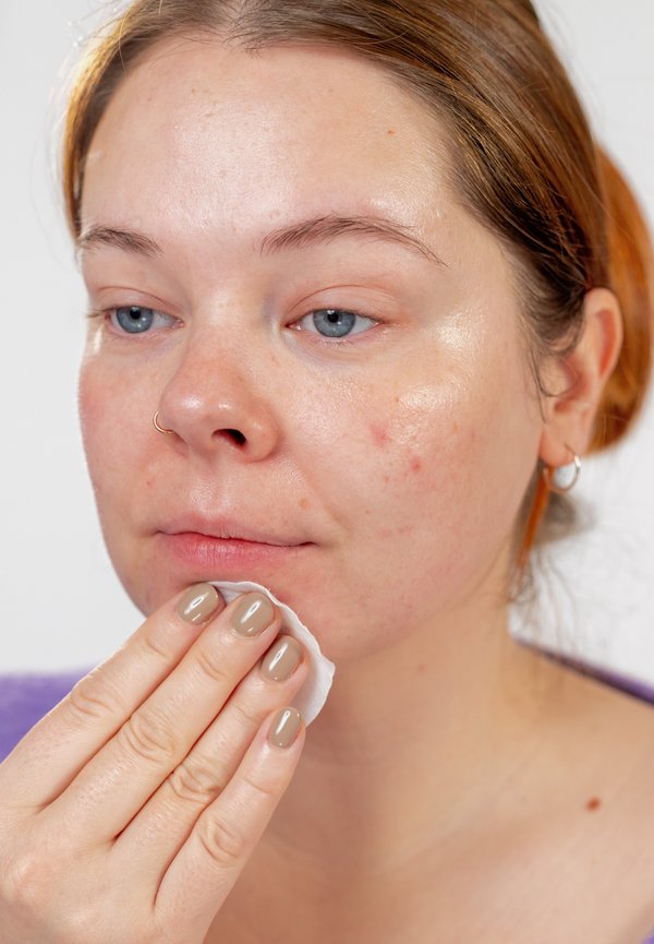 Mujer de piel clara y cabello rojo quitándose el maquillaje o limpiando el rostro con un disco de algodón, mostrando la piel natural con pequeñas imperfecciones.