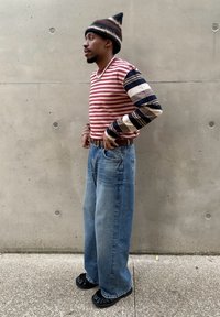 Koszula w paski z długim rękawem w kolorze czerwonym i kremowym, z granatowo-brązowymi wzorzystymi rękawami; luźne niebieskie dżinsy; czarne buty z falbanami; postawa stojąca.