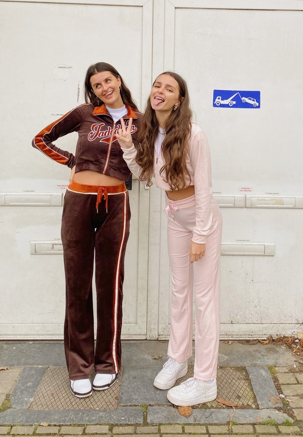 Dos mujeres están de pie contra una pared blanca. Una lleva un chándal de terciopelo marrón con detalles en naranja; la otra lleva un chándal de color rosa claro.