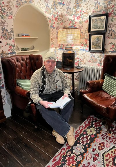 Sillones de cuero con diseño capitoné, papel tapiz estampado, mesa auxiliar de madera y una alfombra texturizada. Persona leyendo un libro, vestida con ropa ligera.