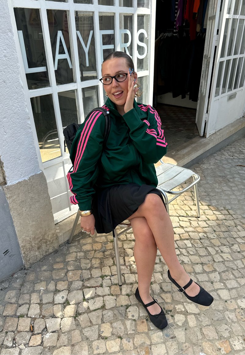Giacca verde con zip e strisce rosa, gonna nera e scarpe Mary Jane nere. Seduta su una sedia di metallo vicino a una vetrina con la scritta "LAYERS."
