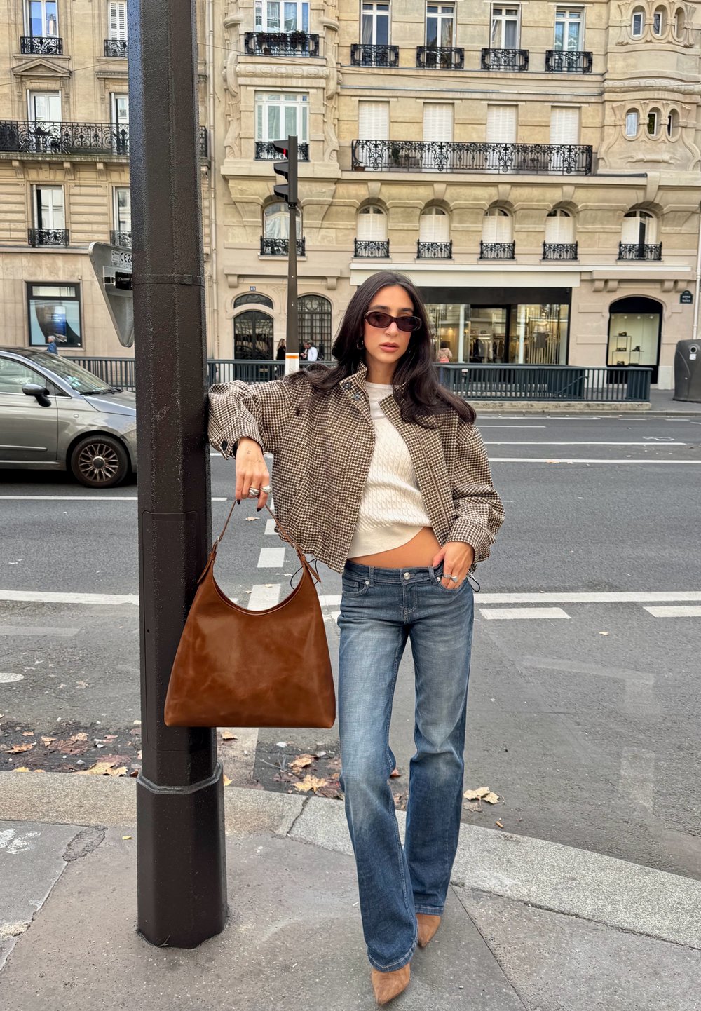 Geantă maro din piele, jachetă cu model houndstooth, bluză albă tricotată, blugi evazați și pantofi cu toc bej. Fundal urban cu clădiri în spate.