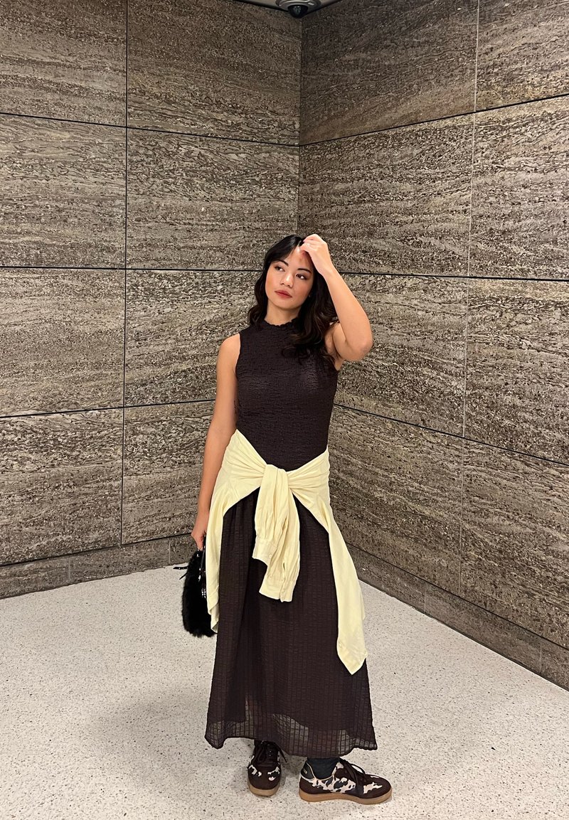 Maxi šaty s texturovaným horním dílem v čokoládově hnědé barvě a splývavou sukní; krémový svetr uvázaný v pase; černá chlupatá kabelka; vzorované boty.