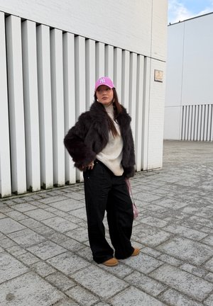 Mujer con gorra rosa, chaqueta de piel negra, suéter beige, pantalones anchos negros y zapatos marrones, de pie en una acera pavimentada cerca de edificios blancos.