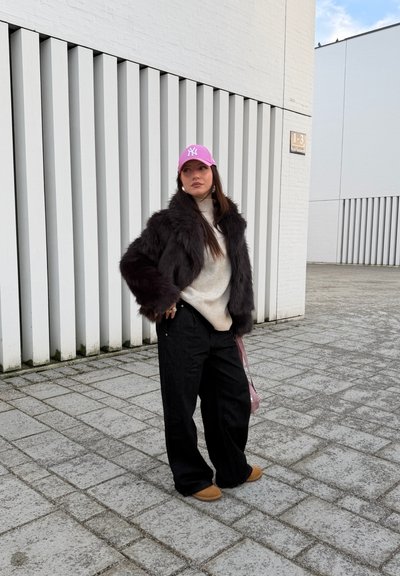Mujer con gorra rosa, chaqueta de piel negra, suéter beige, pantalones anchos negros y zapatos marrones, de pie en una acera pavimentada cerca de edificios blancos.