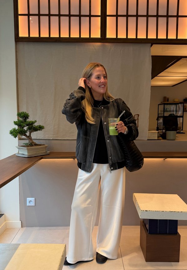 Una mujer con una chaqueta negra y pantalones blancos sostiene una bebida verde en una cafetería moderna, donde hay un bonsái sobre el mostrador.