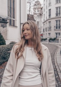 Une femme portant un manteau léger crème sur un haut blanc côtelé, se tenant dans une rue pavée avec des bâtiments en arrière-plan.