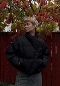 Crna puffer jakna s dijagonalnim patentnim zatvaračem, koja ima visoki ovratnik i oversized kroj. Postavljena na pozadini crvenog i zelenog lišća.