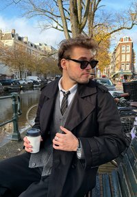 Um homem com um sobretudo preto, suéter cinza, camisa branca e gravata segura uma chávena de café. Usa óculos de sol e está sentado junto a um canal, com bicicletas por perto.