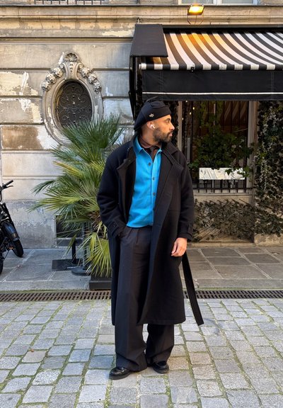 Abrigo negro largo sobre una camisa de botones azul, con pantalones marrones. Zapatos negros y un gorro. Calle de adoquines y una planta de palma en el fondo.