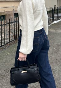 Personne portant un pull blanc et un jean bleu foncé marchant sur un trottoir, tenant un sac à main noir par ses anses dans la main gauche.