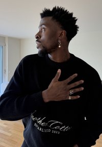 Homme noir aux cheveux courts et bouclés portant un pull noir, des bagues argentées et une boucle d'oreille pendante, regardant vers la gauche avec la main sur la poitrine.