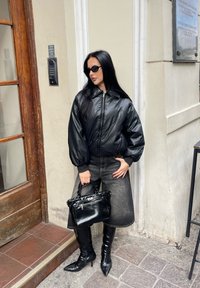 Giacca nera imbottita con colletto alto, abbinata a una gonna di denim scura. Porta una borsa nera e indossa stivali neri al ginocchio.