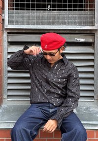 Čierna košeľa so striebornými vertikálnymi pásmi, dlhé rukávy s volánikmi na manžetách, nosená s tmavými džínsami a červenou baretou. Sedí na pozadí z kovu.