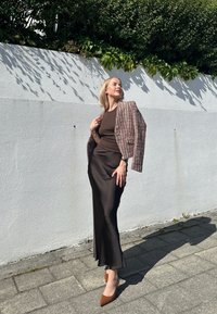 Abito in satin marrone con silhouette aderente, abbinato a un blazer texturizzato multicolore e décolleté marroni a punta, posato su uno sfondo bianco.