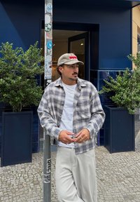 Camisa de franela a cuadros en tonos grises y azules, camiseta blanca debajo, pantalones de color claro. De pie junto a un poste de metal con pegatinas.