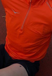 Pomarańczowa sportowa koszulka z półzamkiem, z odblaskowymi akcentami i niebieskim logo. Noszona z czarnymi szortami na teksturowanej powierzchni.