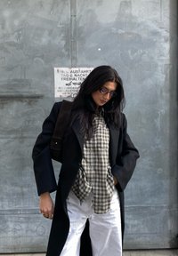 Μαύρο παλτό, καρό πουκάμισο σε μπεζ και σκούρες αποχρώσεις, λευκά παντελόνια και μια σκούρα τσάντα. Μακριά, σκούρα μαλλιά και γυαλιά ηλίου. Γκρίζο φόντο.