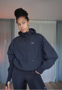 Cortavientos negro de Nike con capucha, ajuste holgado, puños elásticos y bolsillos frontales. Presenta un pequeño logo blanco en el pecho y un diseño minimalista.