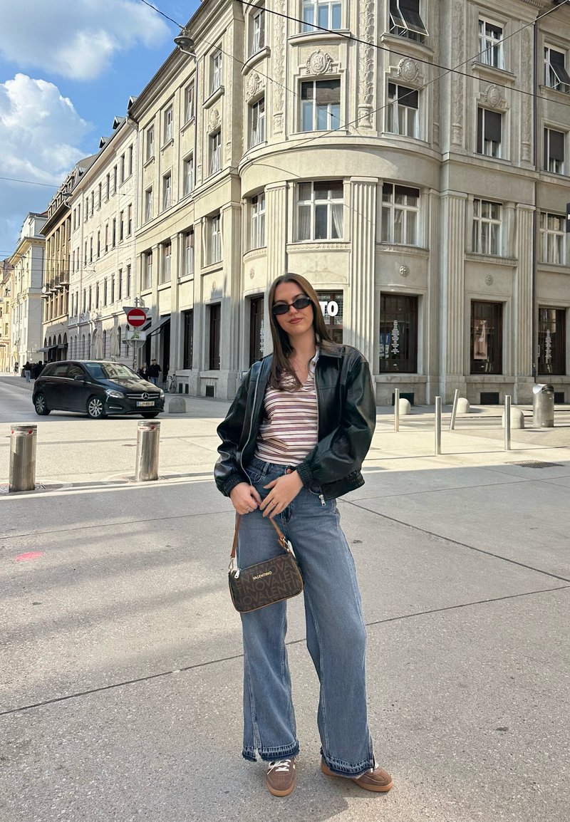 Kvinde iført sort læderjakke, stribet top, vidde jeans og solbriller står på en byhjørne med historiske stenhuse bag sig.