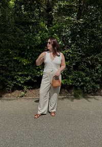 Haut en tricot sans manches de couleur claire avec des boutons ; pantalon large beige ; sac à main en simili cuir marron ; sandales marron ; fond de verdure.