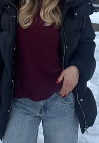 Personne blonde portant une doudoune noire, un pull bordeaux et un jean bleu, avec la main dans la poche, debout dans la neige en plein air.