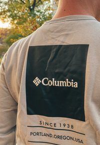 Γκρι μπλούζα με μακριά μανίκια που διαθέτει μια μεγάλη μαύρη γραφιστική απεικόνιση με την ένδειξη "Columbia" και κείμενο από κάτω: "SINCE 1938" και "PORTLAND, OREGON, USA."