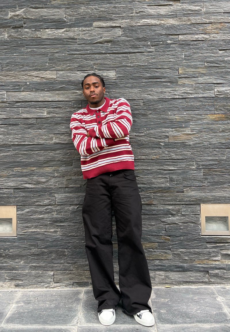Jeune homme avec des tresses portant un pull rayé rouge et blanc, un pantalon noir et des baskets blanches, se tient les bras croisés contre un mur en pierre gris.