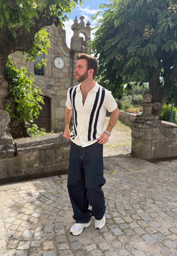 Hombre con barba vestido con camisa blanca con rayas negras y pantalones oscuros, de pie en un camino empedrado cerca de un edificio de piedra con reloj y grandes árboles.