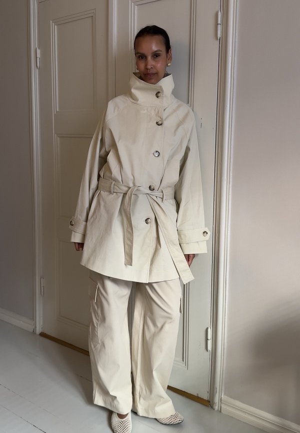Donna in piedi al chiuso che indossa un cappotto color crema con cintura e collo alto, pantaloni a gamba larga abbinati e scarpe bianche perforate.