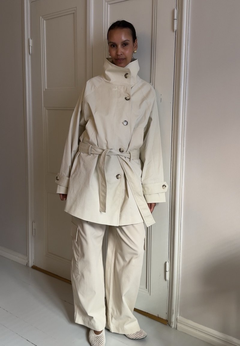 Femme debout à l'intérieur portant un manteau crème ceinturé avec un col montant, un pantalon large assorti, et des chaussures blanches perforées.