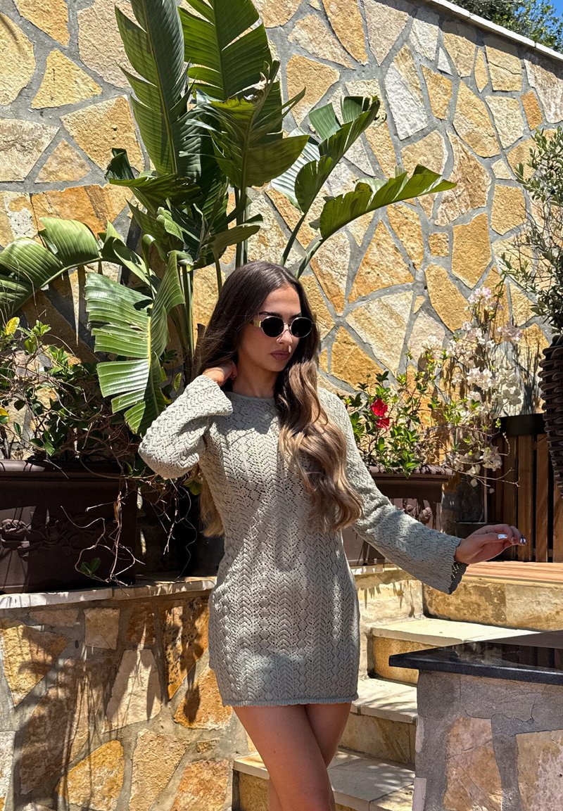 Femme portant une robe en maille grise et des lunettes de soleil posant à l'extérieur près d'un mur en pierre et de grandes plantes vertes, sous un soleil éclatant.