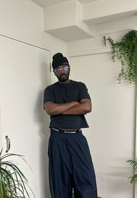 T-shirt noire raccourcie et pantalon bleu marine ample, bonnet noir et lunettes transparentes, avec une plante et un mur blanc en arrière-plan.