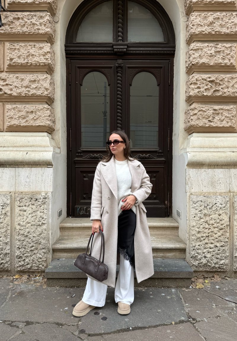 Femme portant un manteau long beige, un pull blanc et un pantalon, une jupe en dentelle noire par-dessus, des chaussures beiges, un sac à main marron, debout devant une porte en bois ornée.