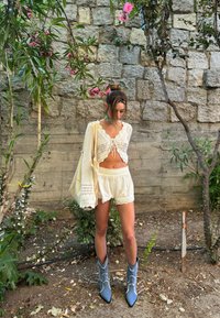 Kvačkana čipkasta kratka majica s vezanjem spredaj, lahki kratki hlače s čipkastim robom in modri kavbojski čevlji z belim šivanjem, v vrtnem ozadju.